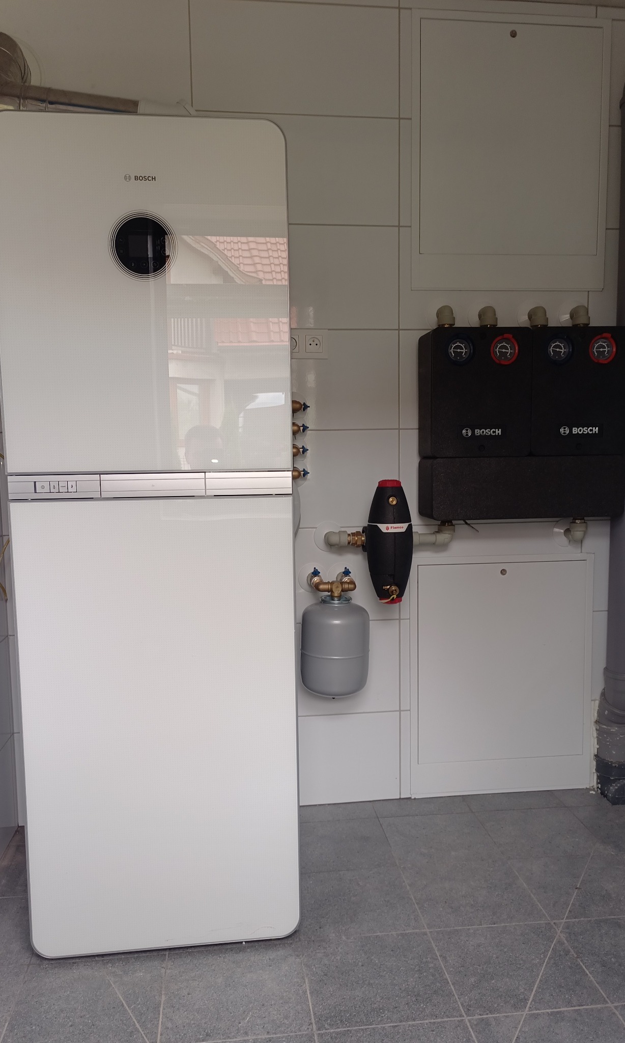 Biały kocioł gazowy Bosch z zasobnikiem i dwoma czarnymi grupami pompowymi Bosch na tle białych płytek, widok z bliska na kotłownię w domu jednorodzinnym.