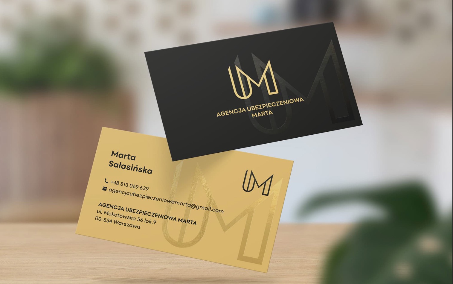 Dwie wizytówki agencji ubezpieczeniowej: złota z danymi kontaktowymi i czarna z logo, obie unoszące się na rozmytym tle biurowym z rośliną.