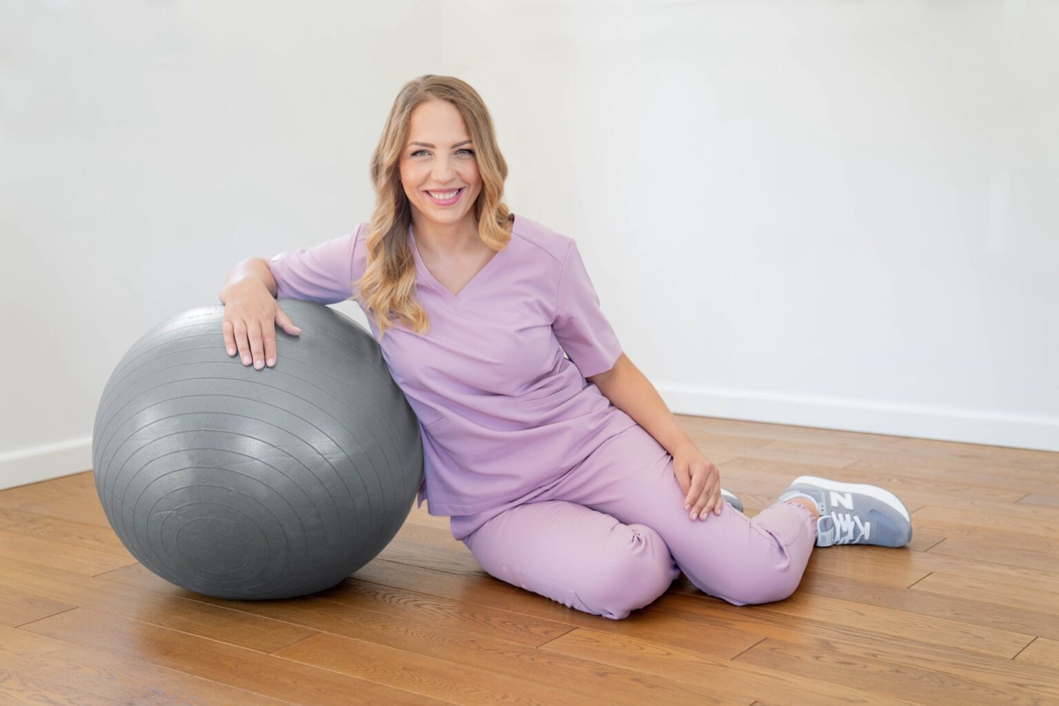 Uśmiechnięta blondynka w liliowym uniformie siedzi oparta o szarą piłkę do ćwiczeń na drewnianej podłodze, w jasnym pomieszczeniu.