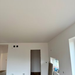 Mateusz Gąciarz - Jasne wnętrze po malowaniu ścian na biało, widoczne gładkie powierzchnie, drewniana podłoga i przygotowane otwory na instalacje elektryczne.
