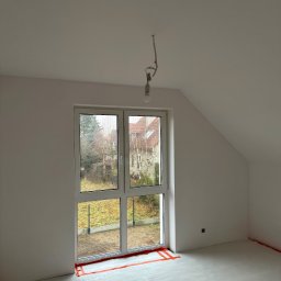 Mateusz Gąciarz - Pokój na poddaszu po szpachlowaniu, przygotowany do malowania. Widoczne zabezpieczenia folią malarską przy oknie i podłodze. Surowa żarówka zwisa z sufitu.