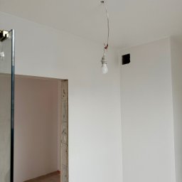 Mateusz Gąciarz - Białe, świeżo pomalowane wnętrze z wiszącą żarówką i widocznym otworem na gniazdko. Widoczny fragment futryny drzwi i surowa podłoga.