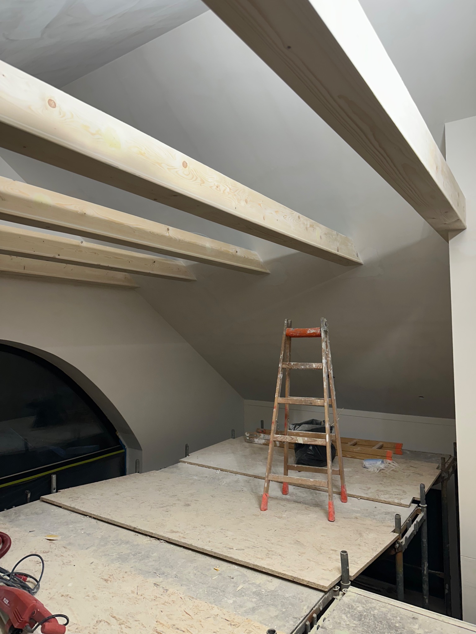 Poddasze w trakcie prac wykończeniowych: drewniane belki, rusztowanie, drabina. Widoczny otwór okienny i narzędzia. Przygotowanie do malowania.
