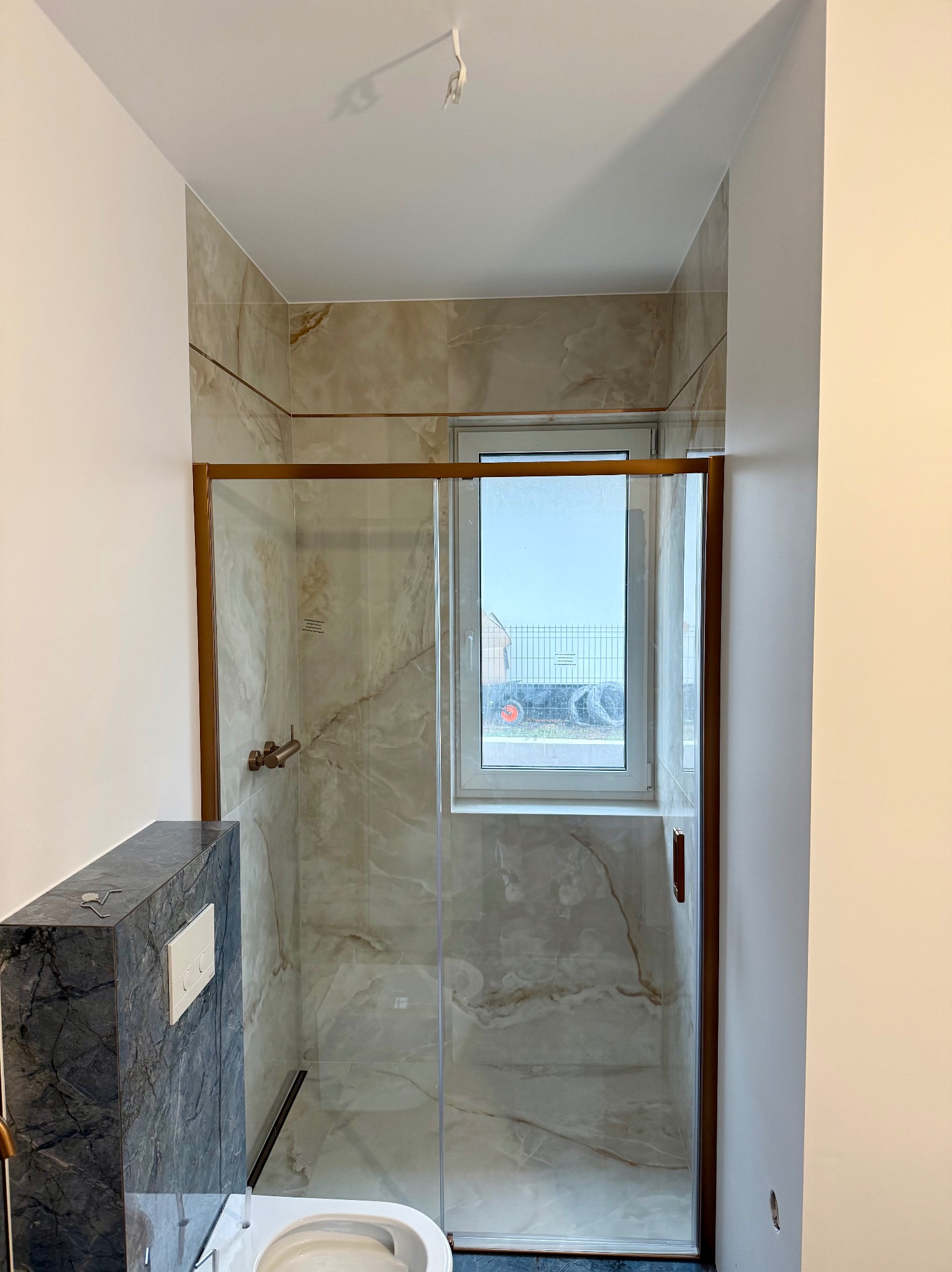 Wykończona łazienka z kabiną prysznicową w odcieniach beżu i złota, marmurowe płytki, widok na okno i fragment toalety. Nowoczesny design.