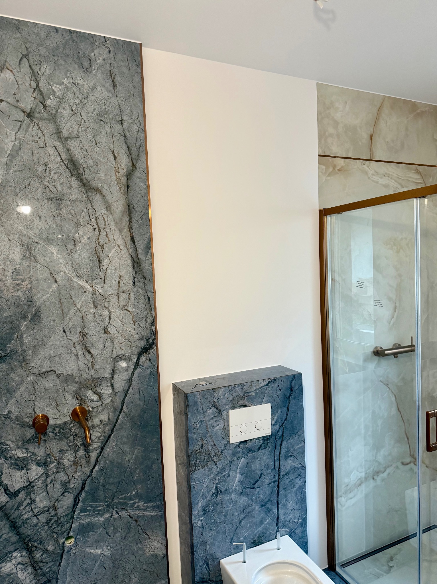 Wykończona łazienka z szarą marmurową ścianą, złotymi detalami armatury i kabiną prysznicową z marmurowym wzorem. Nowoczesny design.