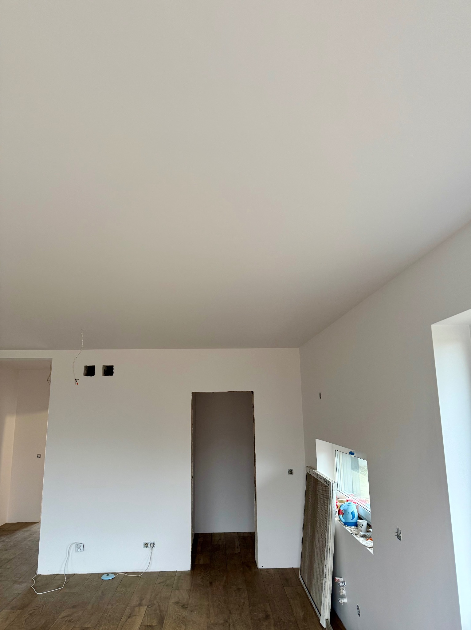 Jasne wnętrze po malowaniu ścian na biało, widoczne gładkie powierzchnie, drewniana podłoga i przygotowane otwory na instalacje elektryczne.