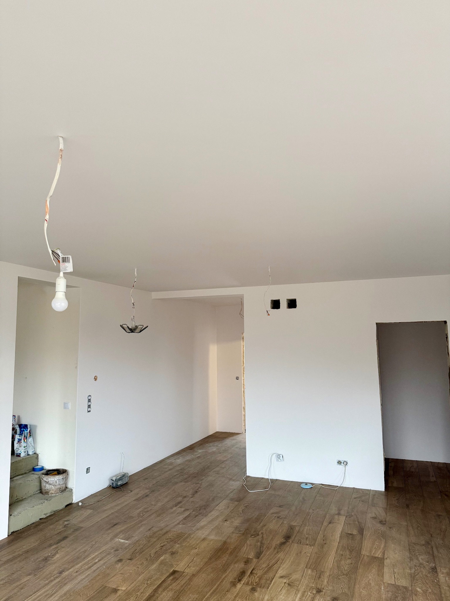 Wnętrze w trakcie remontu: drewniana podłoga, białe ściany z widocznymi puszkami elektrycznymi, wiszące kable i żarówka. Na schodach wiadro i materiały budowlane.