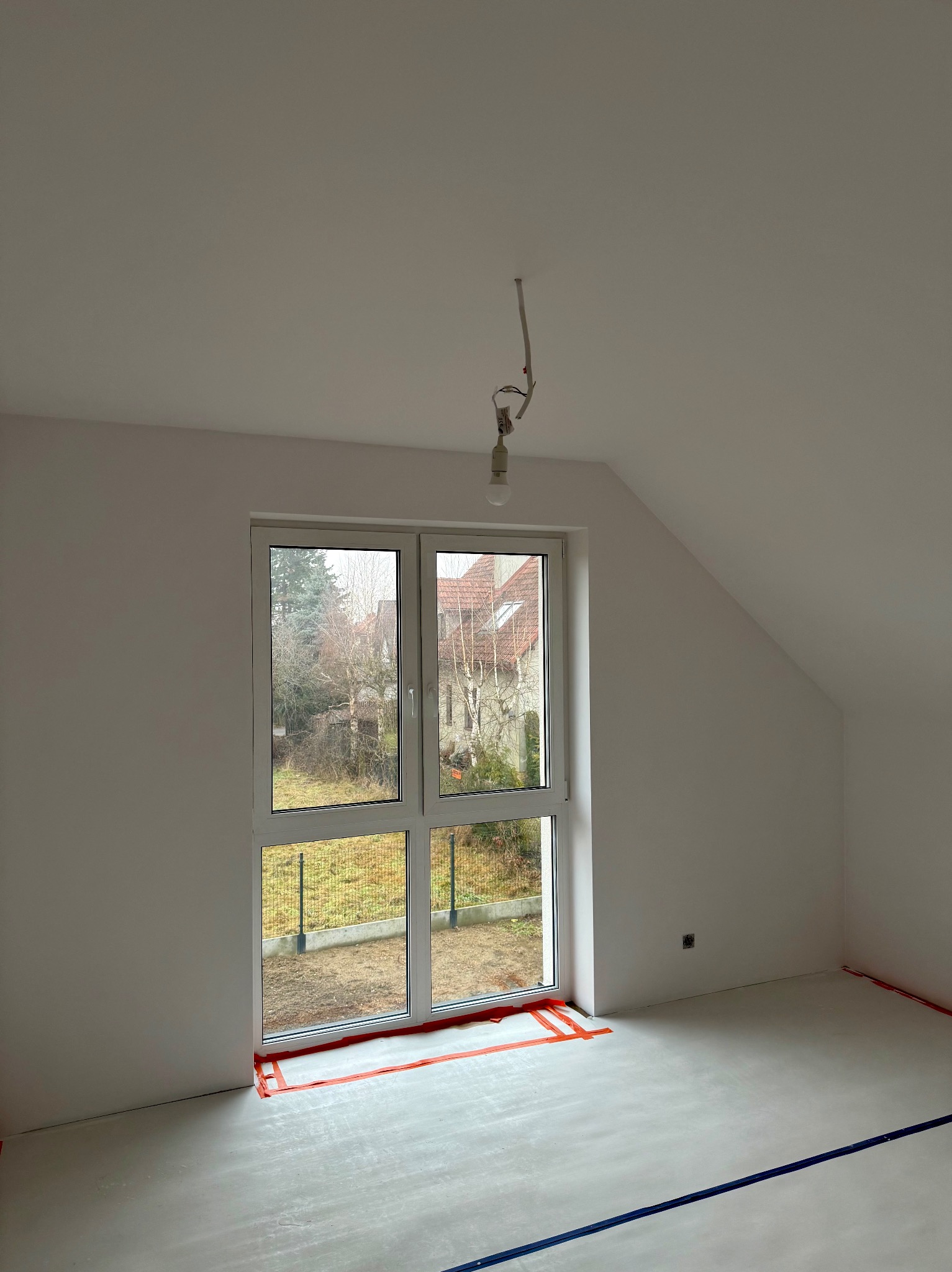 Pokój na poddaszu po szpachlowaniu, przygotowany do malowania. Widoczne zabezpieczenia folią malarską przy oknie i podłodze. Surowa żarówka zwisa z sufitu.