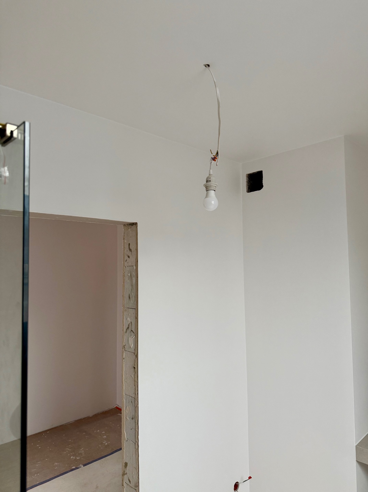 Białe, świeżo pomalowane wnętrze z wiszącą żarówką i widocznym otworem na gniazdko. Widoczny fragment futryny drzwi i surowa podłoga.