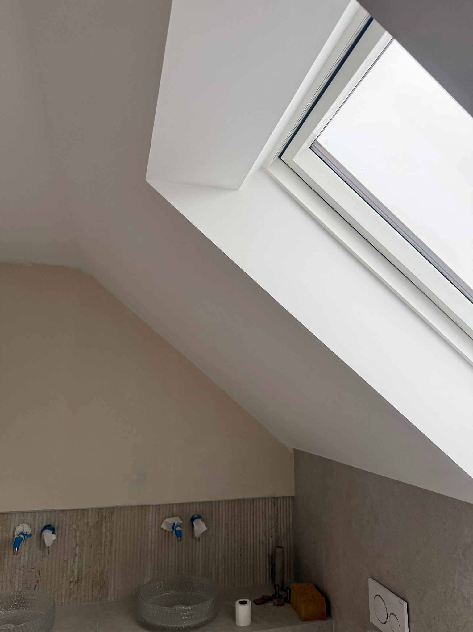 Białe okno dachowe w skosie sufitu, świeżo po malowaniu, z widocznymi detalami wykończenia i fragmentem łazienki z umywalkami. Jasne, naturalne światło.