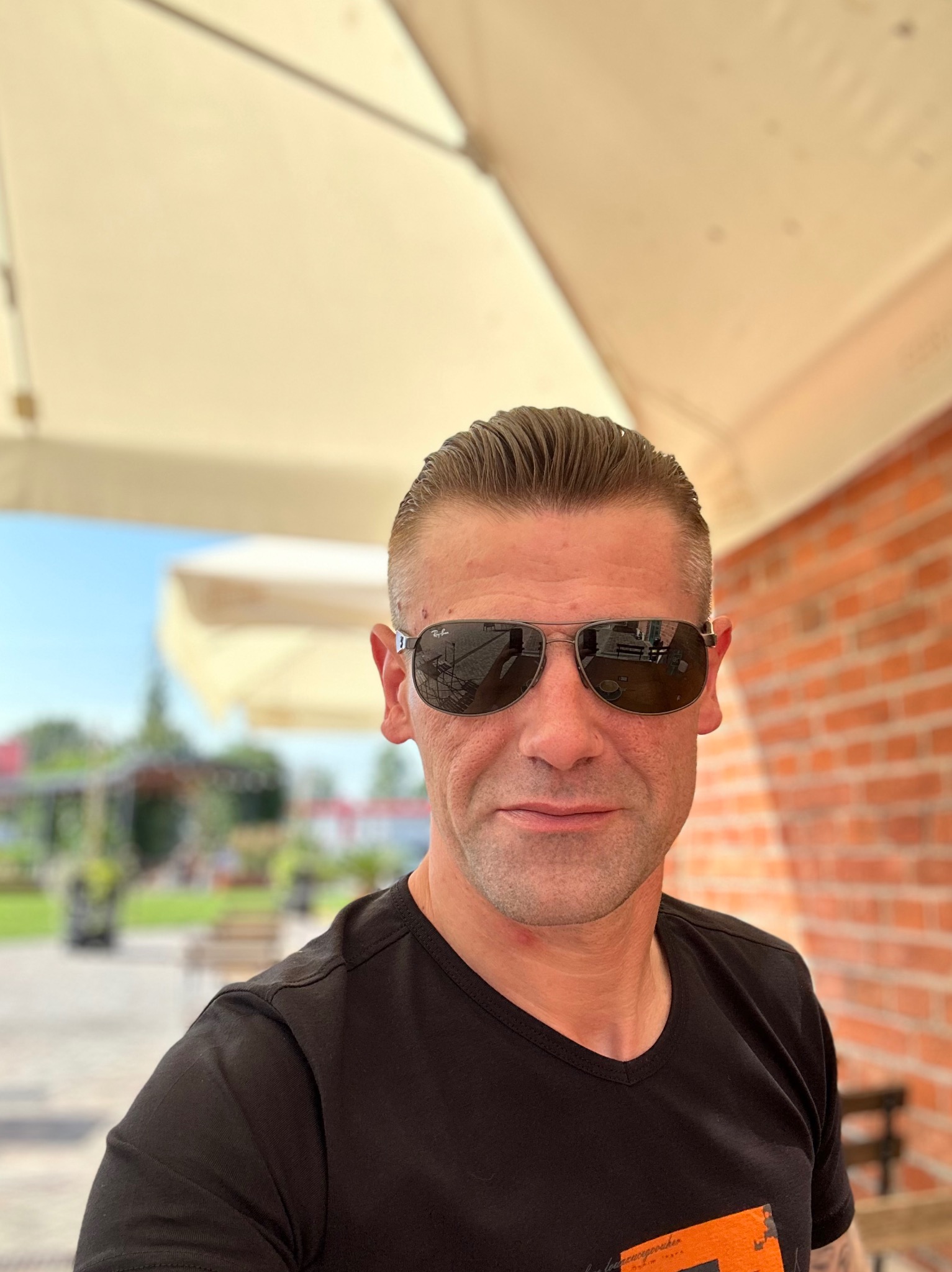 Mężczyzna w okularach przeciwsłonecznych, z ceglaną ścianą w tle, siedzący pod parasolem w plenerowej kawiarni. Widoczne odbicie otoczenia w szkłach okularów.