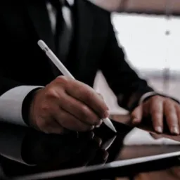 Elegancki mężczyzna w garniturze podpisuje dokument elektroniczny na tablecie za pomocą stylusa. Dyskretne oświetlenie podkreśla profesjonalizm.