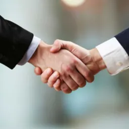 Uścisk dłoni dwóch mężczyzn w garniturach na rozmytym tle, symbolizujący porozumienie lub współpracę biznesową.