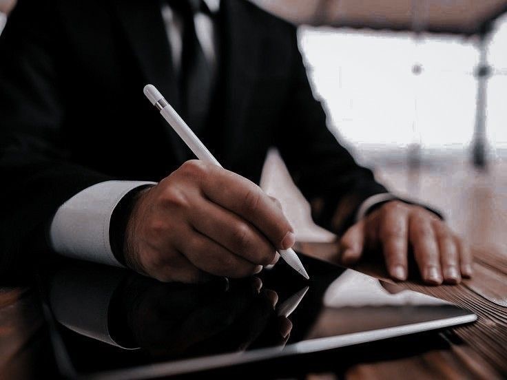 Elegancki mężczyzna w garniturze podpisuje dokument elektroniczny na tablecie za pomocą stylusa. Dyskretne oświetlenie podkreśla profesjonalizm.