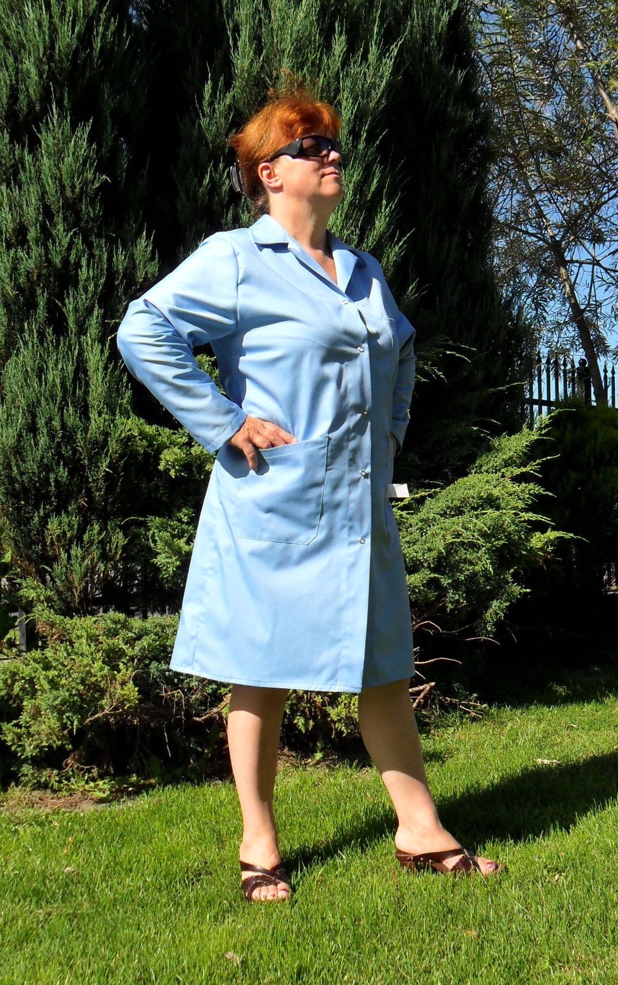 Kobieta w jasnoniebieskim fartuchu medycznym z kieszenią, stojąca na trawie w ogrodzie, w okularach przeciwsłonecznych.