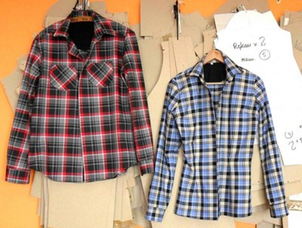 Dwie koszule flanelowe w kratę, czerwono-szaro-czarna z kieszeniami i niebiesko-czarno-biała, wiszące na wieszakach na tle z szablonami krawieckimi.