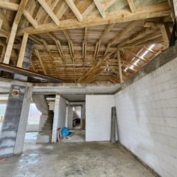 JACEK KRÓLCZYK STELLARIS - Wnętrze w stanie surowym z drewnianą konstrukcją dachu, widoczne białe ściany z bloczków, betonowa podłoga i schody. Etap budowy domu.