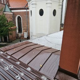 JACEK KRÓLCZYK STELLARIS - Nowy, brązowy dach z blachy na budynku z cegły z wieżyczką. Widoczne mocowania i ślady po deszczu na dachu. Komin z prawej strony kadru.