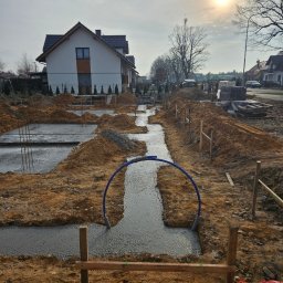 JACEK KRÓLCZYK STELLARIS - Wylane fundamenty pod budowę domu jednorodzinnego, widoczne szalunki i rury instalacyjne. W tle domy w zabudowie jednorodzinnej i błękitne niebo.