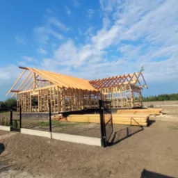 Konstrukcja drewniana domu szkieletowego w trakcie budowy, widok zza ogrodzenia. Na pierwszym planie stos drewna, w tle błękitne niebo z chmurami.