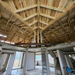 JACEK KRÓLCZYK STELLARIS - Wnętrze surowego domu w trakcie budowy: betonowe kolumny, ściany z białych bloczków i drewniana konstrukcja dachu z widocznymi belkami.