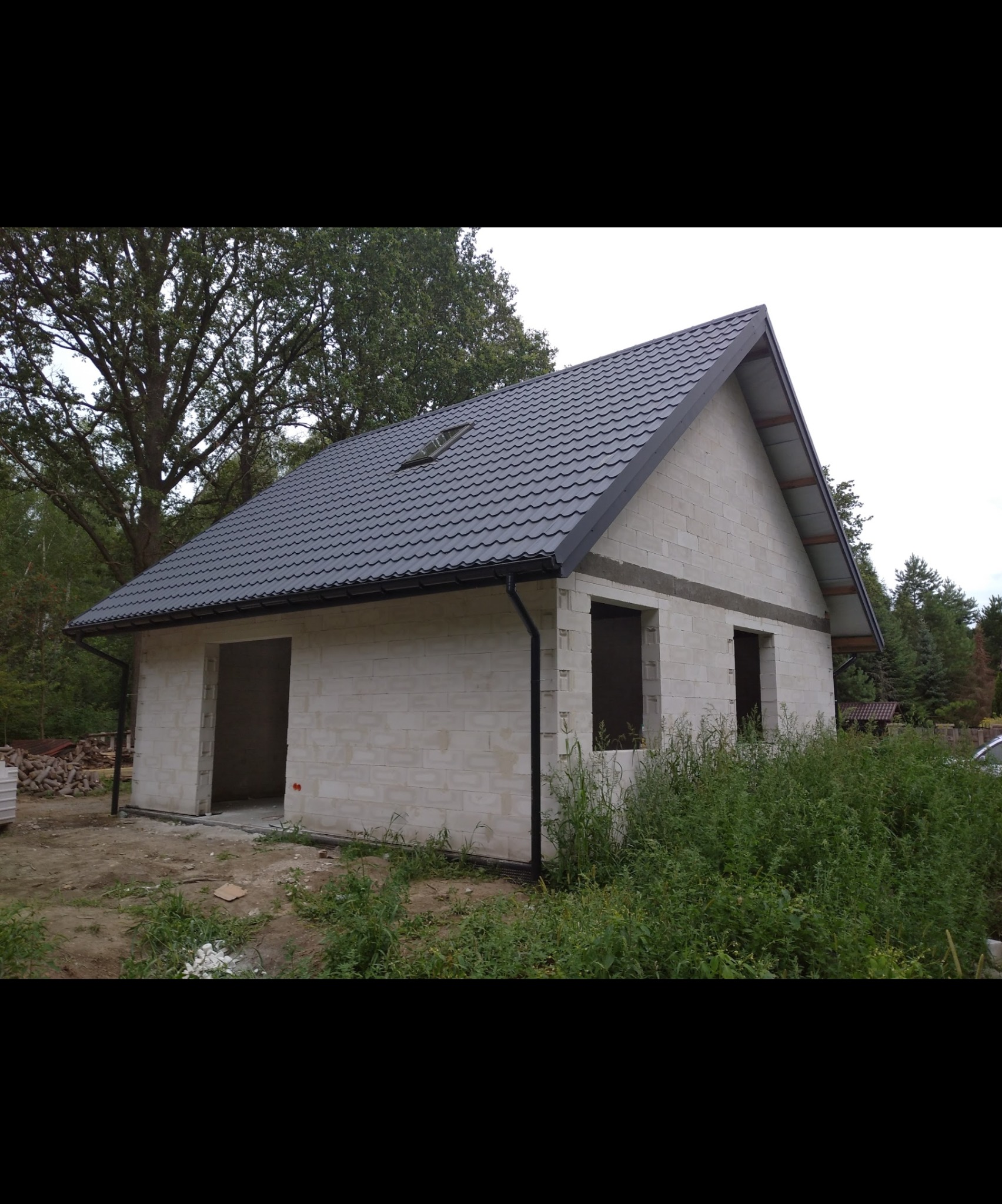 Niedokończony dom z bloczków, z szarą dachówką i orynnowaniem. Widoczne otwory okienne i drzwiowe, teren wokół budynku częściowo zagospodarowany zielenią.