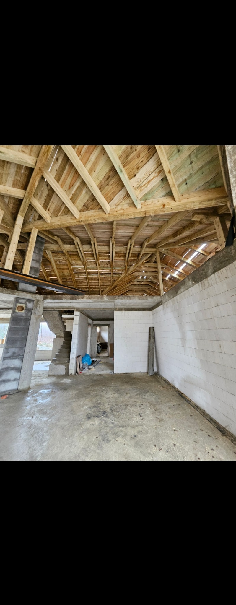 Wnętrze w stanie surowym z drewnianą konstrukcją dachu, widoczne białe ściany z bloczków, betonowa podłoga i schody. Etap budowy domu.