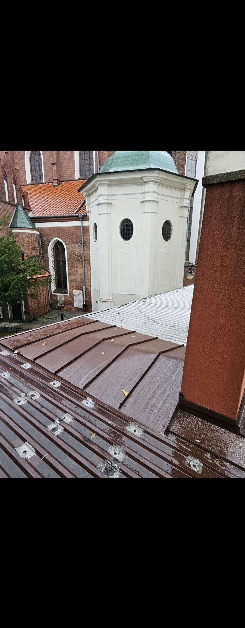 Nowy, brązowy dach z blachy na budynku z cegły z wieżyczką. Widoczne mocowania i ślady po deszczu na dachu. Komin z prawej strony kadru.