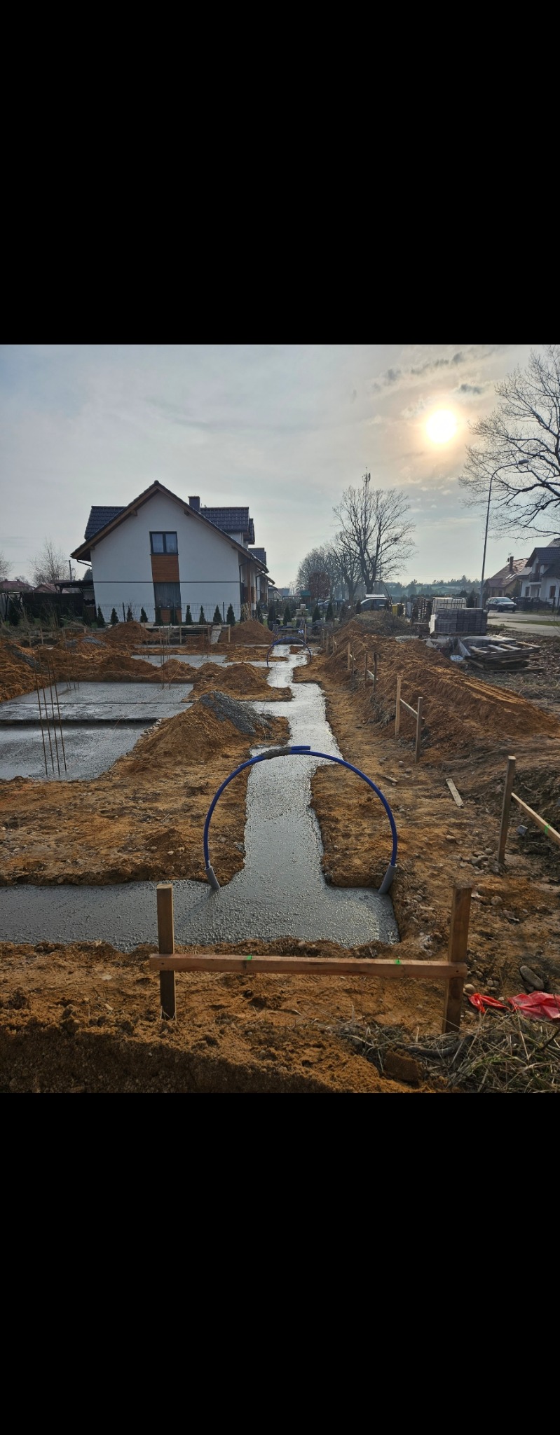 Wylane fundamenty pod budowę domu jednorodzinnego, widoczne szalunki i rury instalacyjne. W tle domy w zabudowie jednorodzinnej i błękitne niebo.