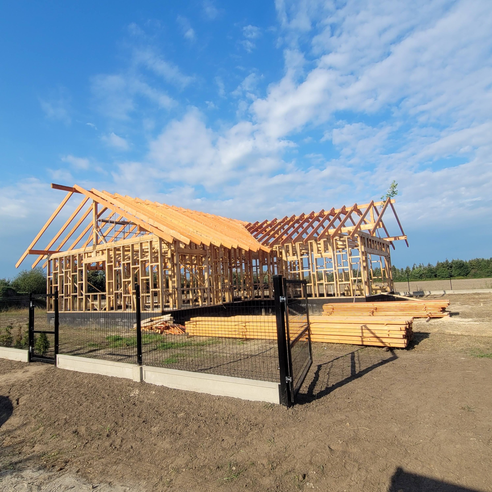Konstrukcja drewniana domu szkieletowego w trakcie budowy, widok zza ogrodzenia. Na pierwszym planie stos drewna, w tle błękitne niebo z chmurami.