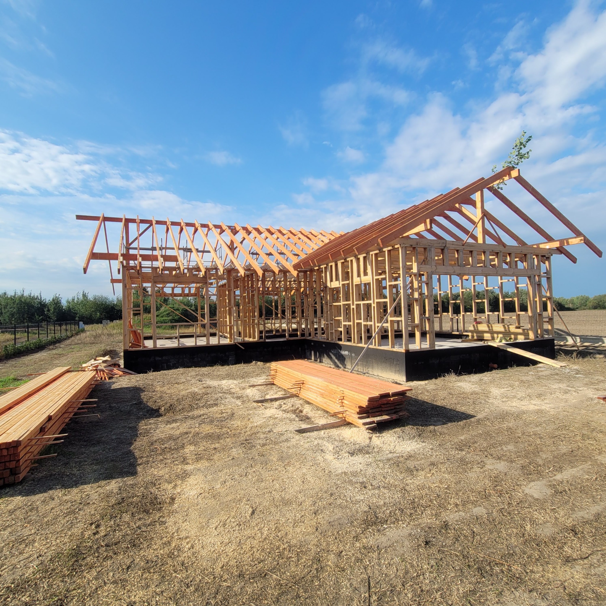 Drewniana konstrukcja domu w trakcie budowy, widok z zewnątrz, na pierwszym planie deski, błękitne niebo w tle. Etap wznoszenia ścian i dachu.