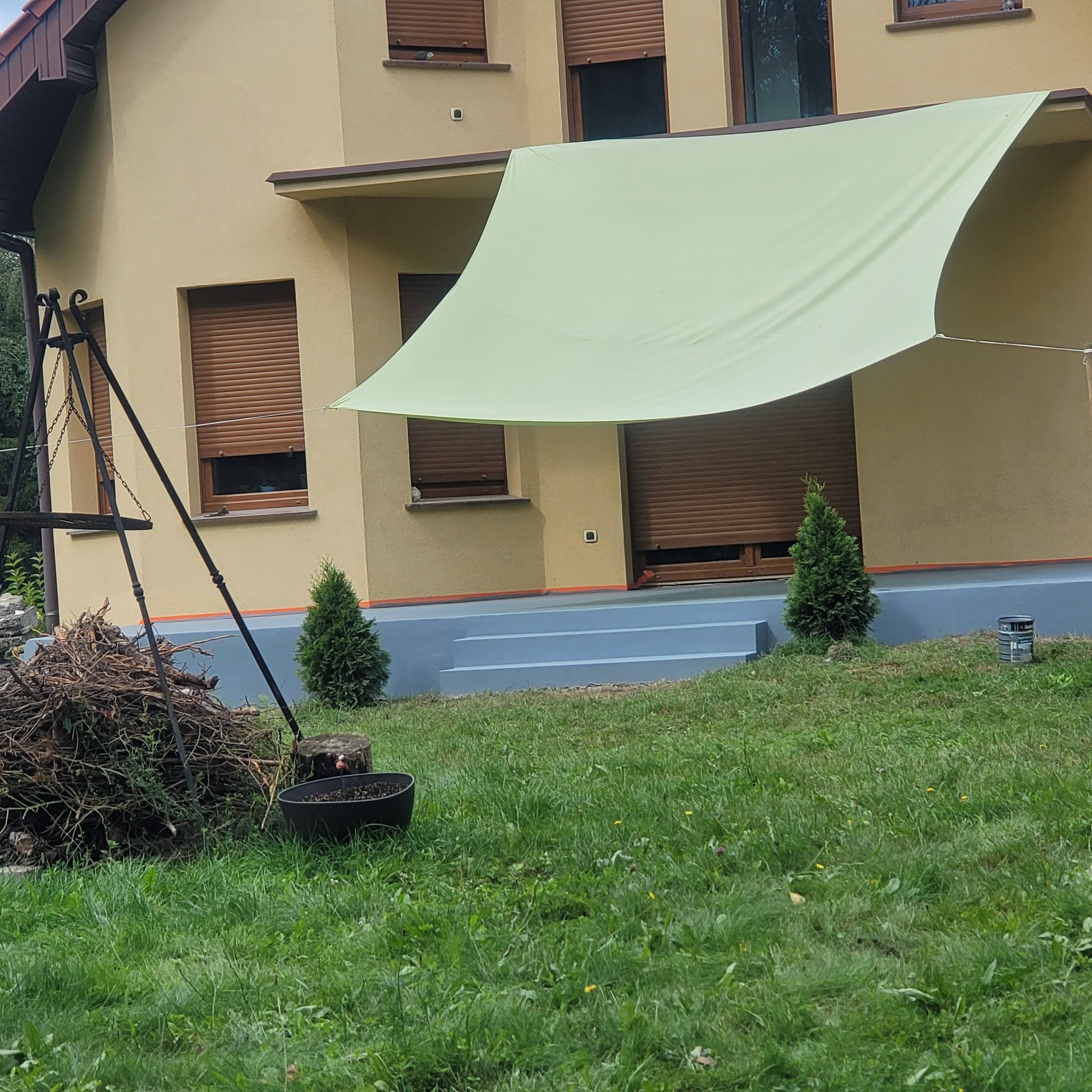 Beżowy dom z brązowymi roletami i zieloną markizą przeciwsłoneczną. Widoczne pomalowane schody wejściowe na szaro i dekoracyjne drzewka iglaste.