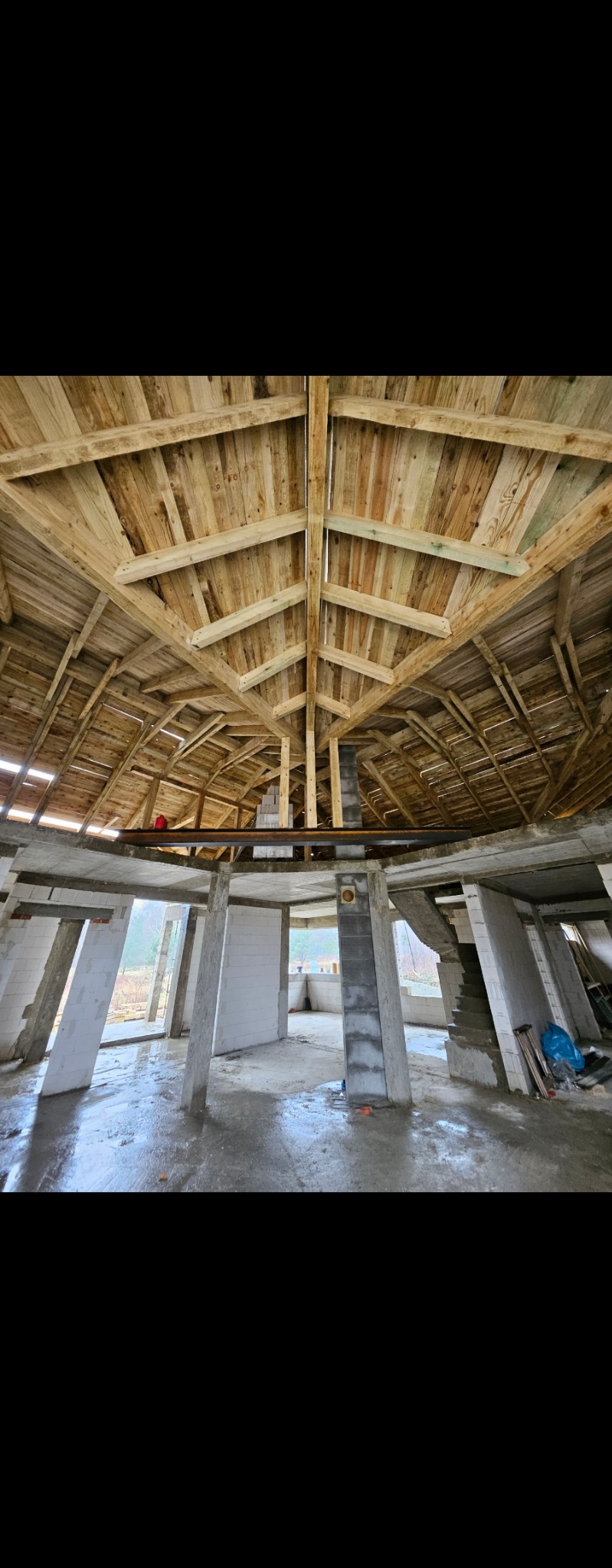 Wnętrze surowego domu w trakcie budowy: betonowe kolumny, ściany z białych bloczków i drewniana konstrukcja dachu z widocznymi belkami.