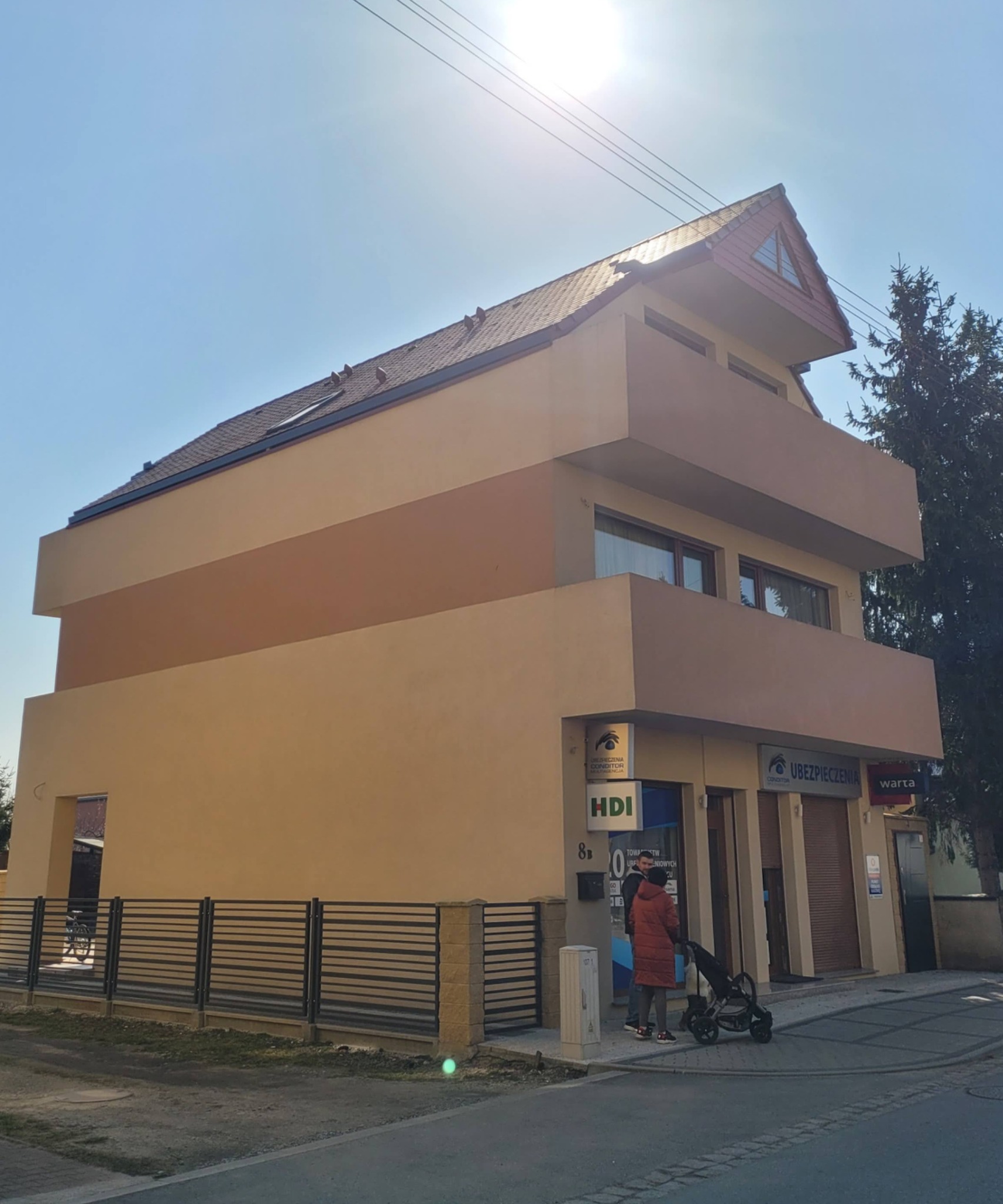 Dwukondygnacyjny budynek o nietypowej bryle z balkonami i oknami, pomalowany w odcieniach beżu i brązu, z widocznymi szyldami firm ubezpieczeniowych na parterze.