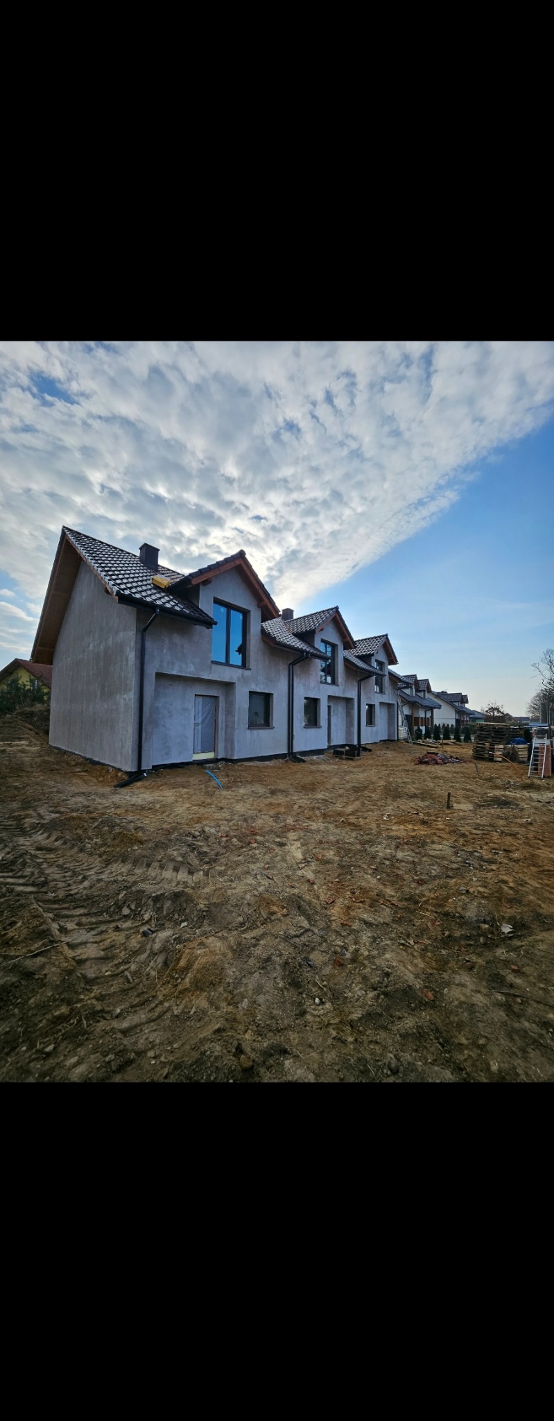 Szereg surowych, otynkowanych domów jednorodzinnych z ciemną dachówką, widocznych z poziomu gruntu, na tle błękitnego nieba z chmurami. Etap budowy.