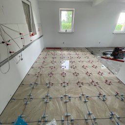 RemontFactory - Układanie beżowych płytek podłogowych z systemem poziomowania w jasnym pomieszczeniu. Częściowo wykończona podłoga, widoczne narzędzia i worki z klejem.