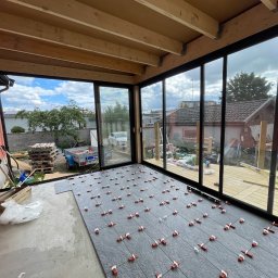 RemontFactory - Układanie szarych płytek podłogowych z dystansami w przeszklonym pomieszczeniu z drewnianym stropem. Widoczne narzędzia i częściowo ułożona podłoga.