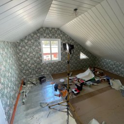 RemontFactory - Poddasze z oknem po tapetowaniu ścian w kwiatowy wzór. Na podłodze folia ochronna i narzędzia, lampa na statywie oświetla wnętrze. Widoczne resztki tapet.