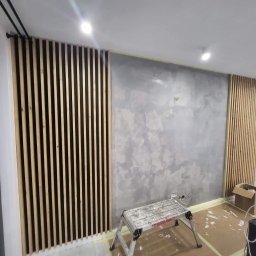 RemontFactory - Szara ściana z efektem dekoracyjnym i drewniane lamele, fragment drabiny malarskiej. Wnętrze w trakcie wykańczania.