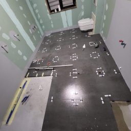 RemontFactory - Układanie ciemnych płytek podłogowych w łazience, z białymi krzyżykami dystansowymi. Częściowo przygotowane ściany z płyt karton-gips. Widoczne narzędzia glazurnicze.