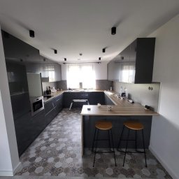 RemontFactory - Nowoczesna kuchnia w odcieniach szarości z błyszczącymi frontami szafek, drewnianym blatem i dekoracyjną podłogą. Widoczne dwa stołki barowe i drabinka.