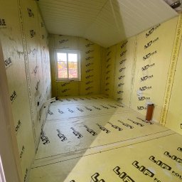 RemontFactory - Wnętrze w trakcie prac wykończeniowych: ściany i podłoga pokryte żółtą folią izolacyjną z czarnym nadrukiem, widoczne okno i fragment sufitu.