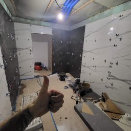 RemontFactory - Układanie płytek w łazience z użyciem krzyżyków dystansowych. Widoczna ręka z kciukiem w górę. W tle narzędzia glazurnicze i częściowo ułożone płytki imitujące marmur.