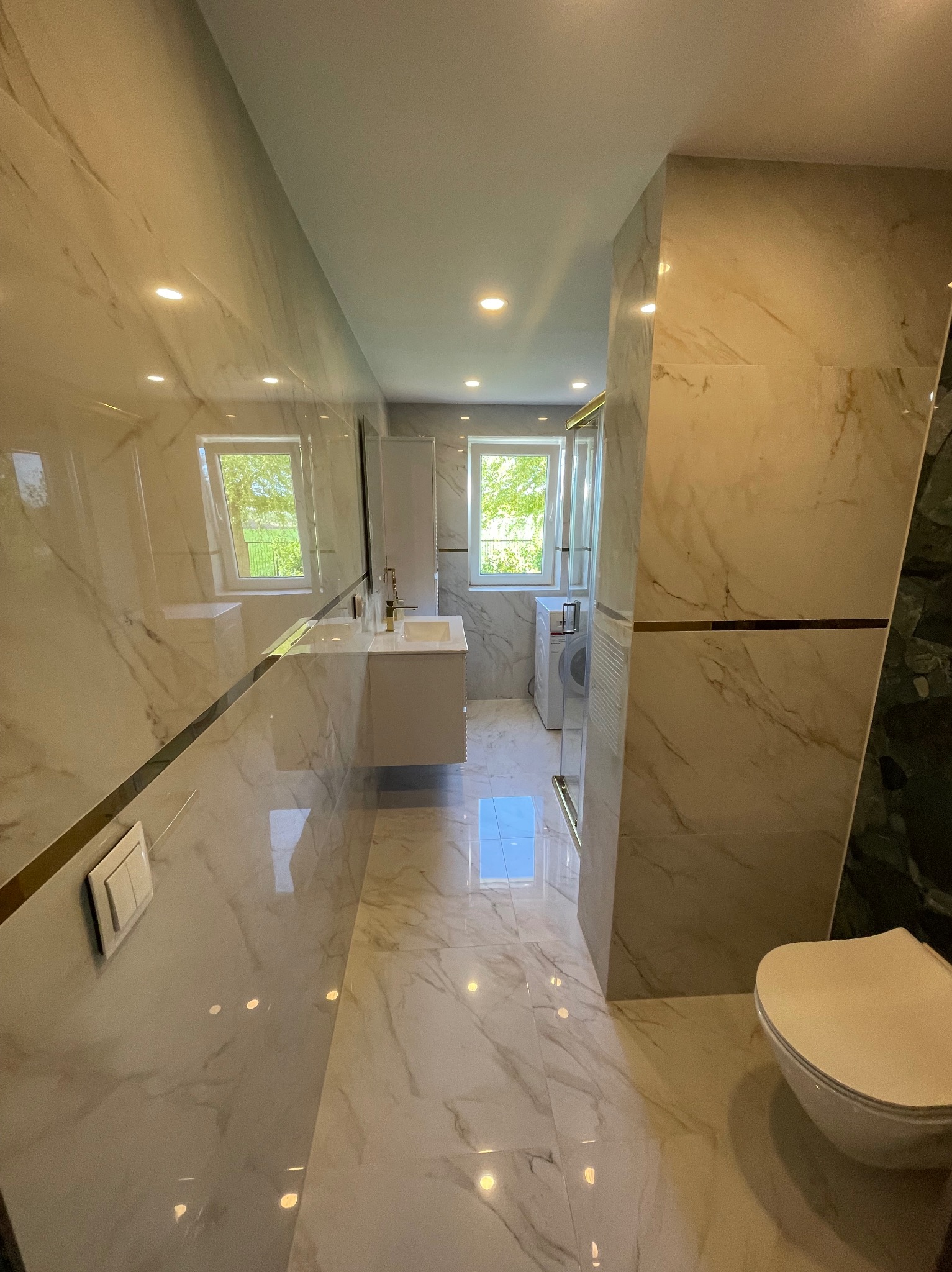 Jasna łazienka z marmurowymi płytkami, złotymi akcentami i dużym lustrem, odbijającym okno. Widoczne umywalka, toaleta i kabina prysznicowa.