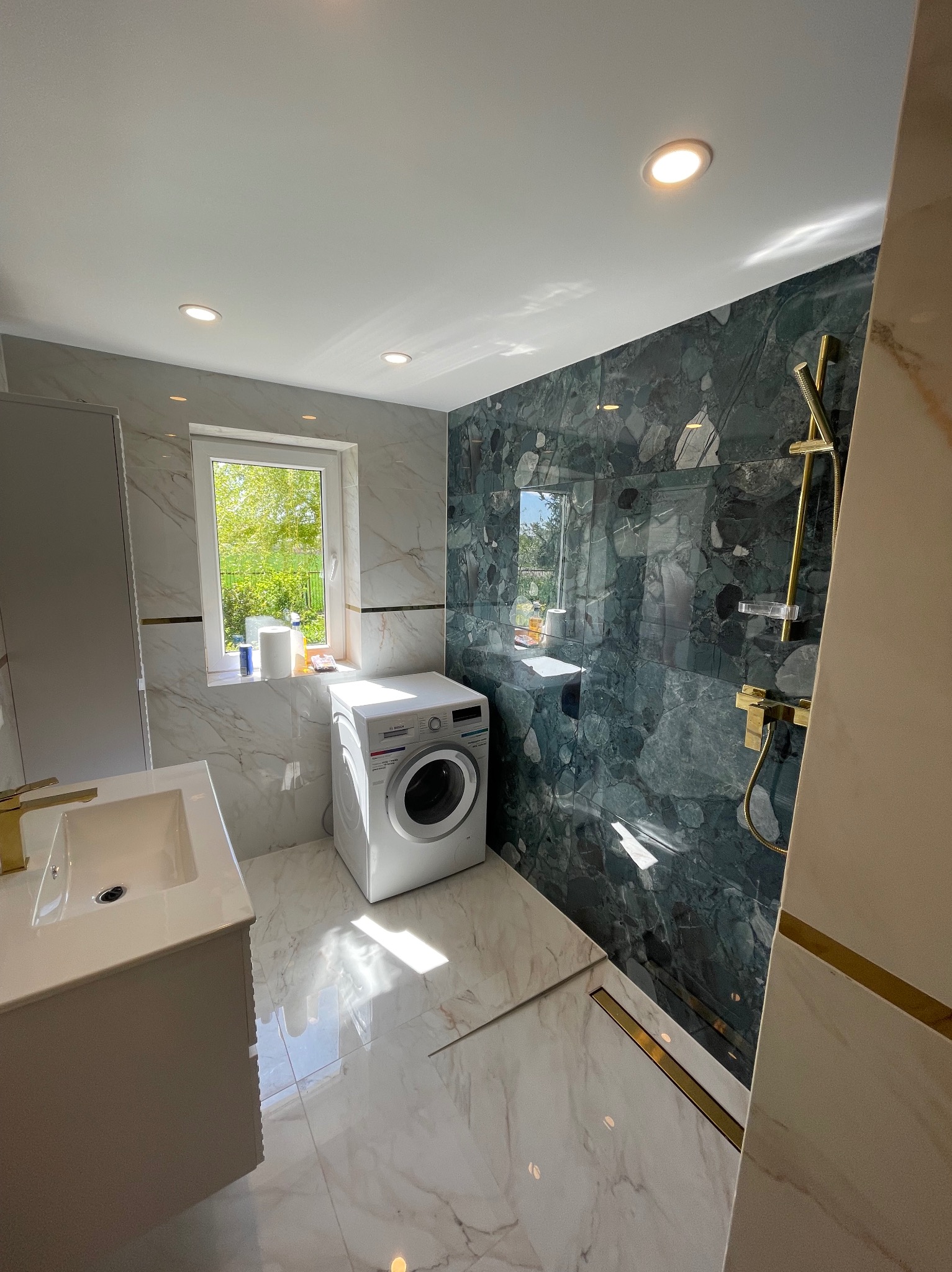 Wykończona łazienka w bieli i zieleni z złotymi akcentami: marmurowe płytki, umywalka z baterią, pralka Bosch pod oknem, kabina prysznicowa z deszczownicą.