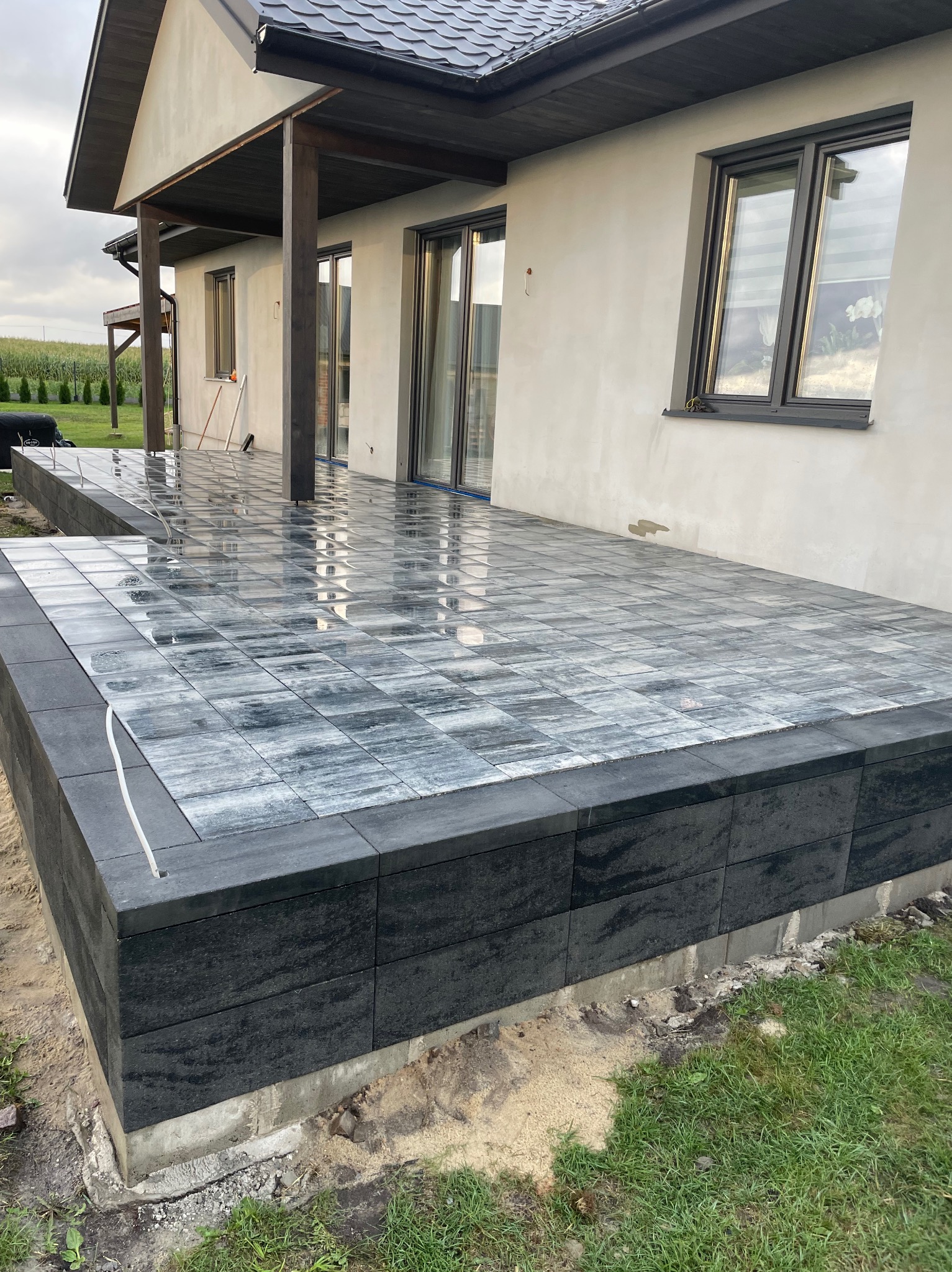 Ułożona kostka brukowa na tarasie domu jednorodzinnego w szaro-czarnej tonacji. Widoczne okna i fragment elewacji. Kostka mokra, odbija światło.