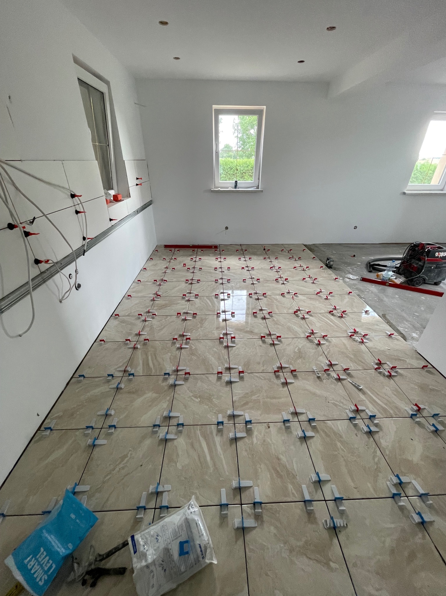 Układanie beżowych płytek podłogowych z systemem poziomowania w jasnym pomieszczeniu. Częściowo wykończona podłoga, widoczne narzędzia i worki z klejem.