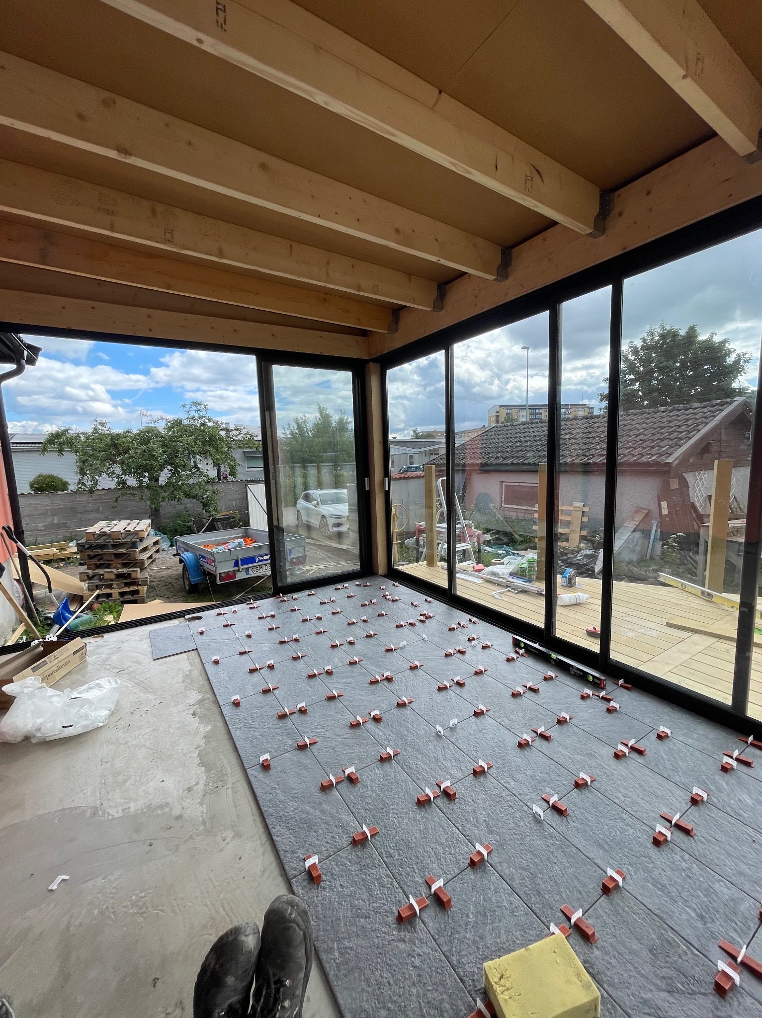 Układanie szarych płytek podłogowych z dystansami w przeszklonym pomieszczeniu z drewnianym stropem. Widoczne narzędzia i częściowo ułożona podłoga.