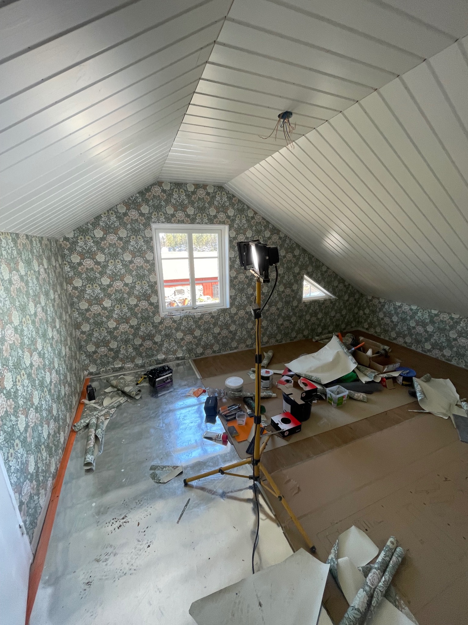 Poddasze z oknem po tapetowaniu ścian w kwiatowy wzór. Na podłodze folia ochronna i narzędzia, lampa na statywie oświetla wnętrze. Widoczne resztki tapet.
