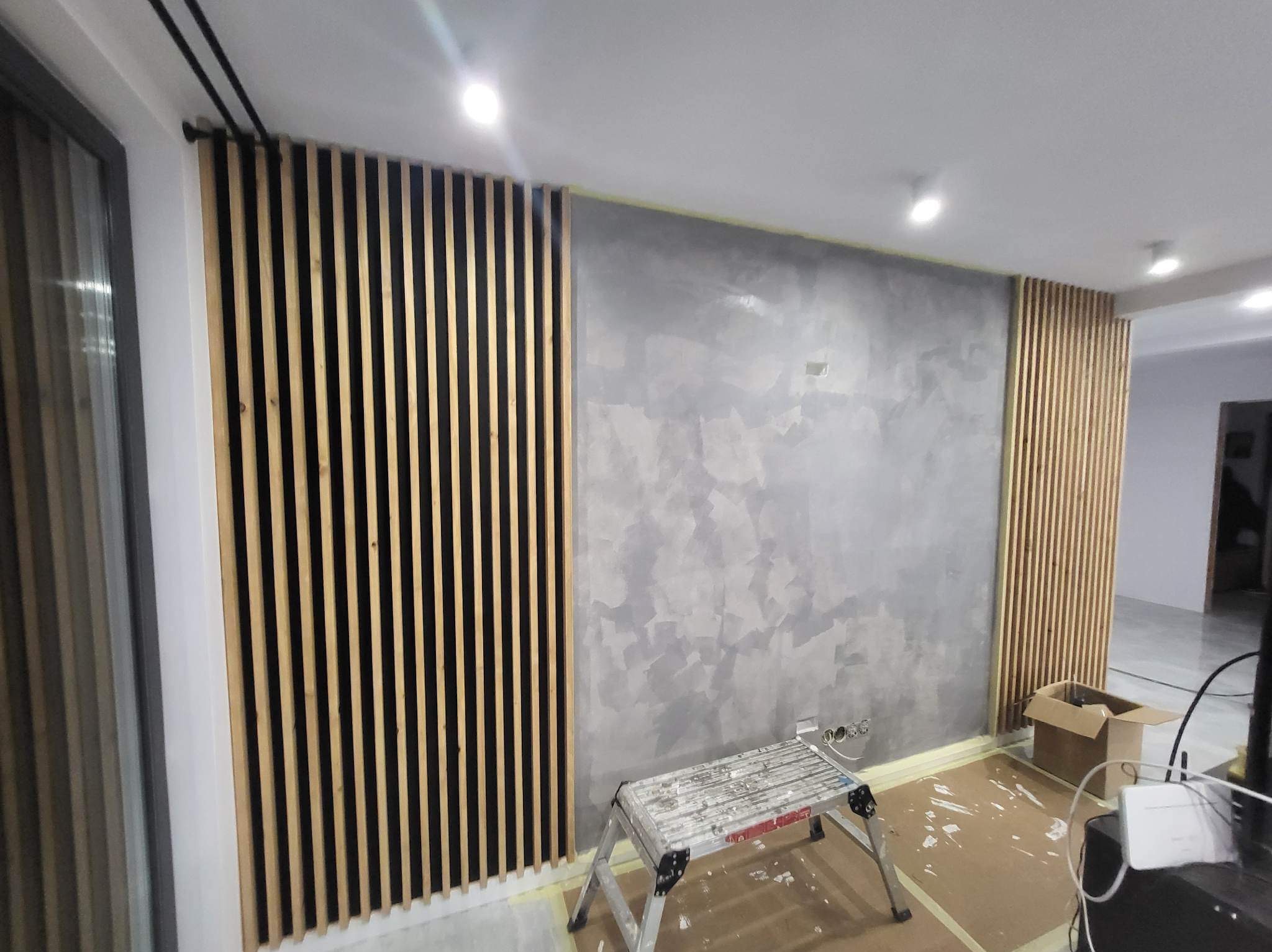 Szara ściana z efektem dekoracyjnym i drewniane lamele, fragment drabiny malarskiej. Wnętrze w trakcie wykańczania.